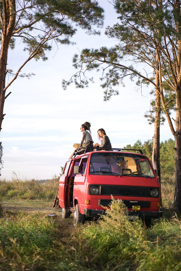 team-04 Two women enjoy a scenic view sitting on a red van during a road trip through lush greenery.