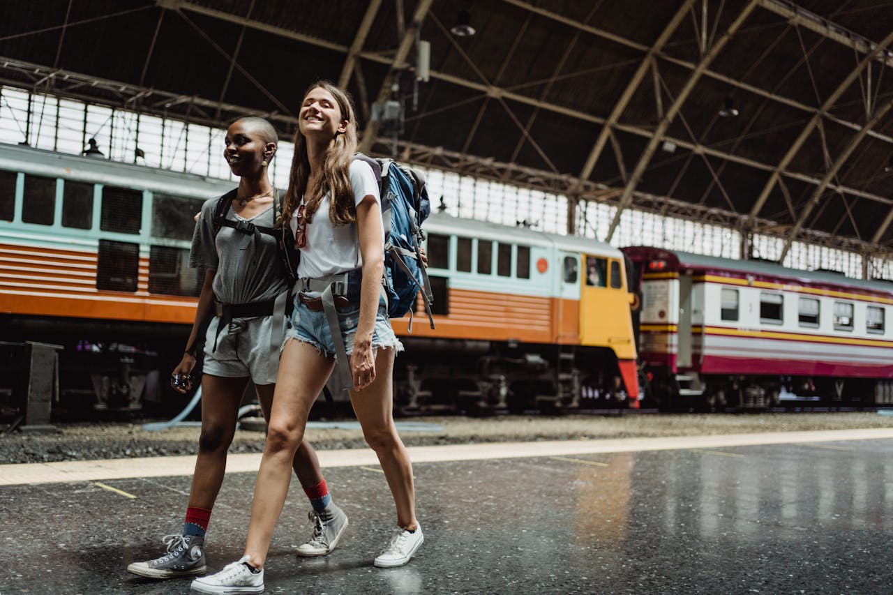 team-03 Two women with backpacks walking and smiling at a railway station.